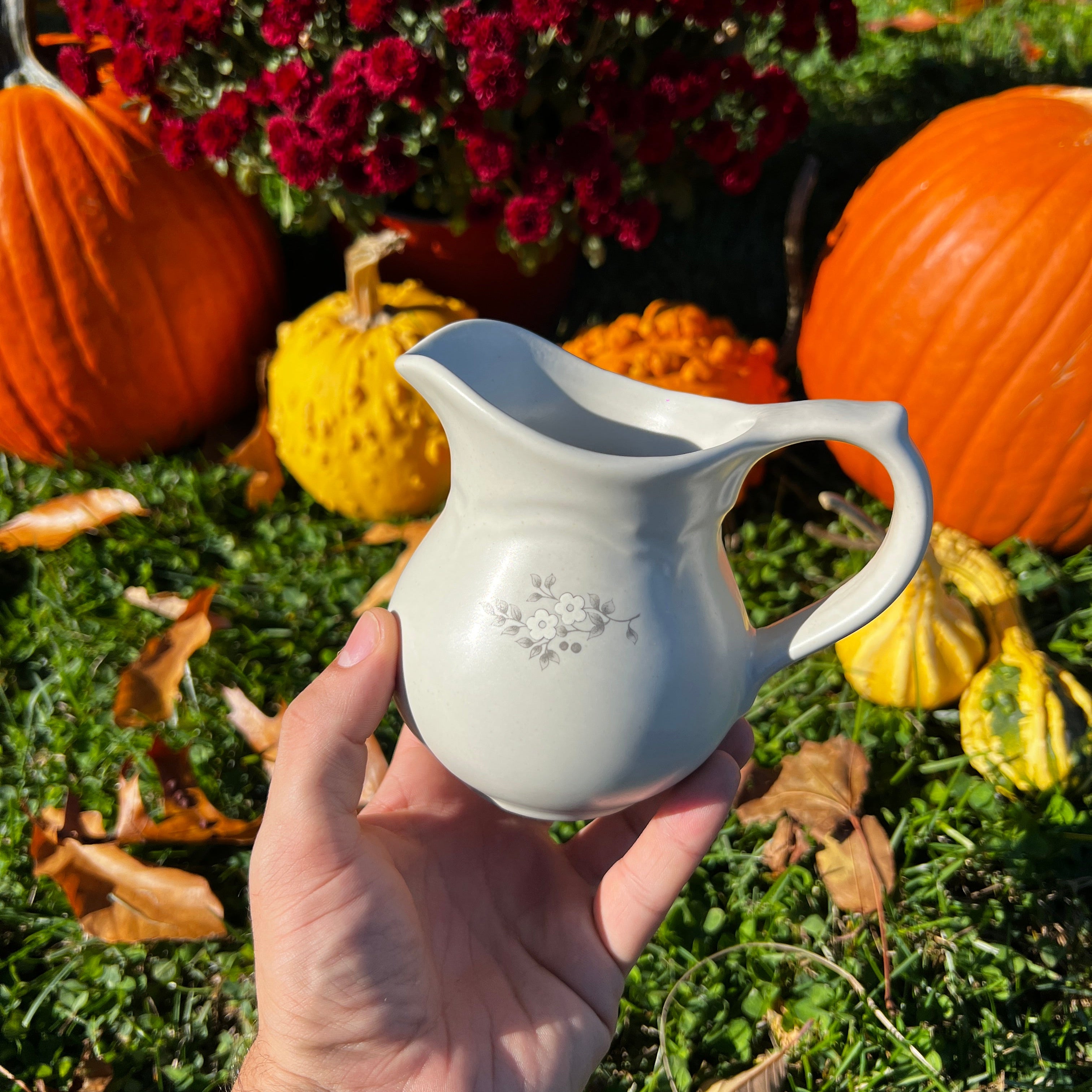 Hand holding a white ceramic pitcher with pumpkins and flowers in the background